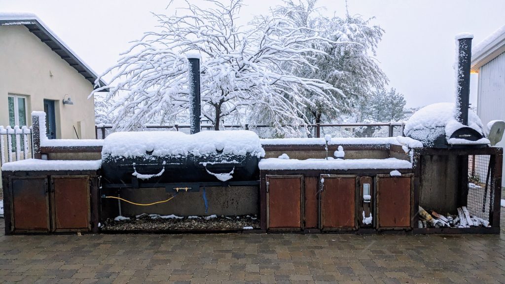 Black BBS smoker and metal pizza oven covered in snow.